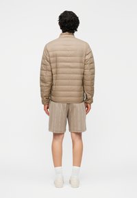 Bézs színű steppelt pufi dzseki vízszintes steppeléssel, magas gallérral és könnyű anyagból, bézs mintás rövidnadrággal és fehér zoknival párosítva.