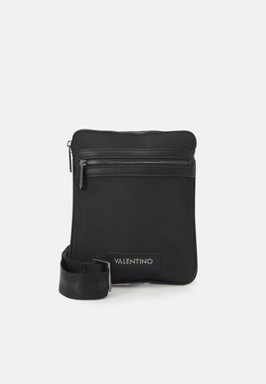 Zwarte Valentino crossbodytas met voorvak met ritssluiting, verstelbare riem en logopatch op een witte achtergrond.