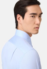 Camicia azzurra chiara con colletto strutturato, texture liscia e design aderente. Presenta una cucitura della spalla dritta e una pattina con bottoni.