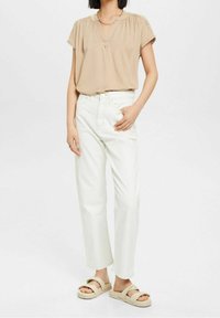 Blouse beige à manches courtes avec un décolleté en V, associée à un jean blanc coupe droite et des sandales slides beiges avec une semelle épaisse.