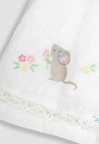 Tela blanca con un ratón bordado en marrón sosteniendo una flor, acompañado de flores en tonos pastel. Un borde de encaje en la parte inferior añade un detalle.