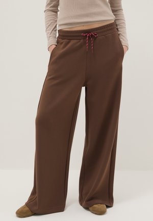 Persona con pantalones marrones de pierna ancha y cintura ajustable con cordón, camiseta beige de manga larga y zapatos marrones, de pie con las manos en los bolsillos.