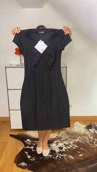 Pessoa atrás de um vestido preto até aos joelhos com decote em V e mangas curtas, segurando-o pelos ombros, em pé sobre um tapete de pele de animal castanho e branco.