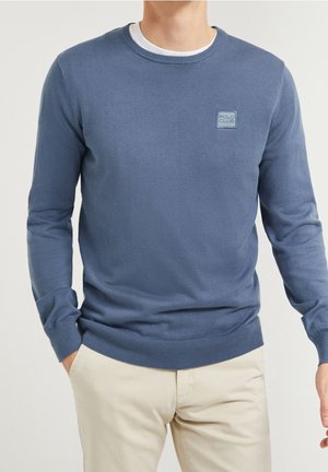 Blaues Langarmoberteil aus weichem Stoff, mit Rundhalsausschnitt und kleinem quadratischen Logo auf der Brust. Glatte Textur.