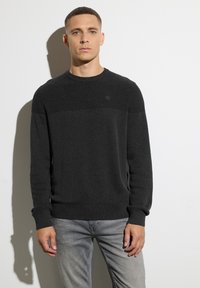 Dunkelgrauer Strickpullover mit strukturiertem Oberteil, Rundhalsausschnitt und Markenlogo auf der Brust. Slim-Fit, kombiniert mit hellgrauen Jeans.