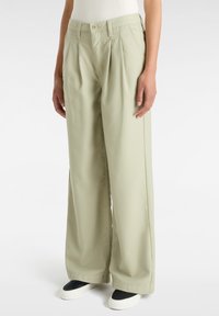 Pantaloni larghi verde chiaro con vita alta, caratterizzati da pinces frontali e una consistenza liscia. Indossati con sneakers bianche e nere.