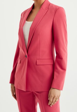 Vrouw draagt een op maat gemaakt koraalroze pantalonpak met een blazer met één knoop over een witte geribbelde top, staand met één zichtbare hand met een ring aan de vinger.