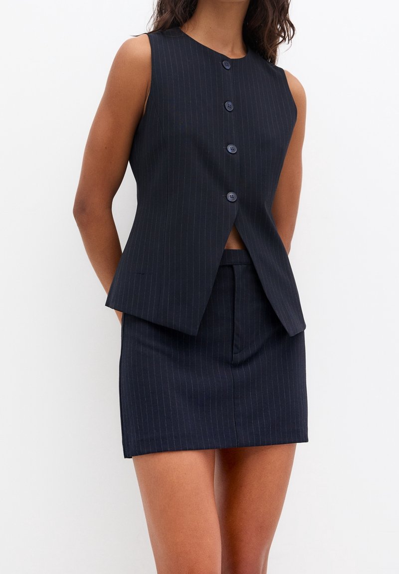 Gilet sans manches bleu marine à fines rayures avec boutons, associé à une mini-jupe taille haute assortie, porté par une personne debout.