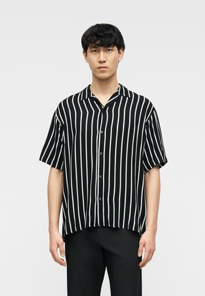 Junger Mann mit kurzen schwarzen Haaren, der ein schwarzes Kurzarm-Hemd mit weißen vertikalen Streifen und schwarze Hose trägt.