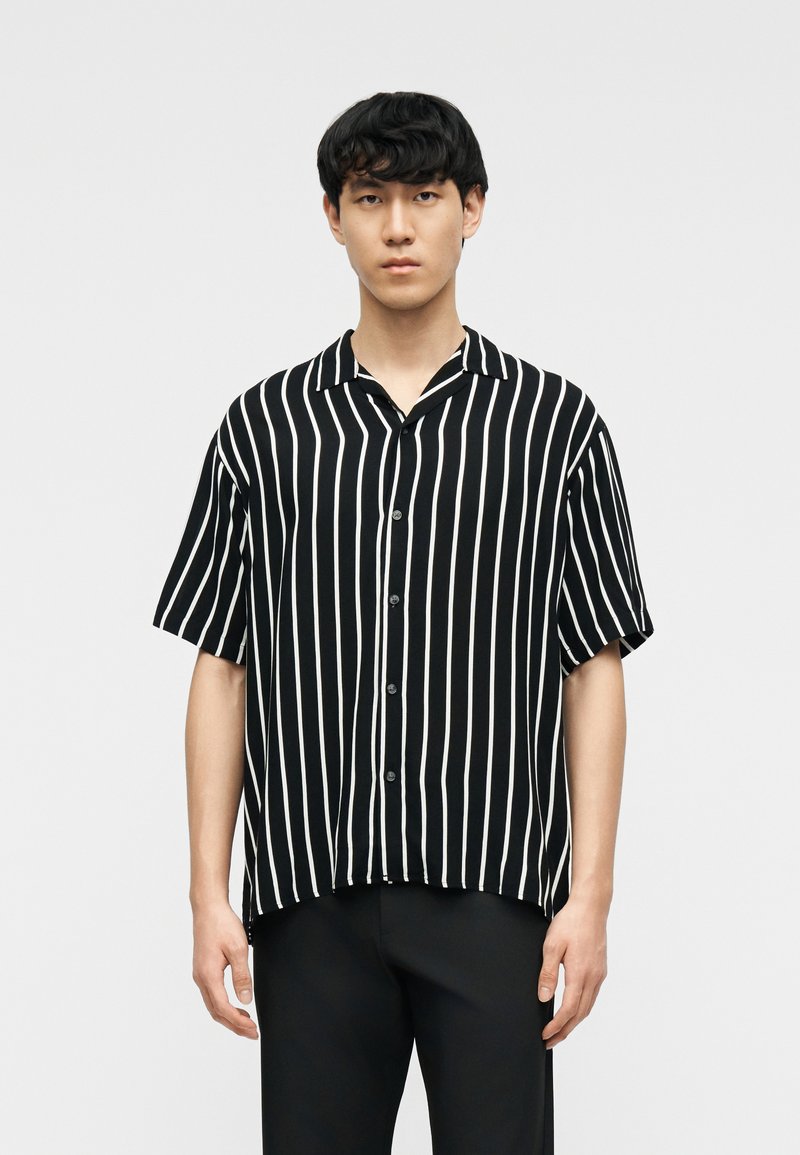 Νέος άνδρας με κοντά μαύρα μαλλιά, που φοράει μαύρο πουκάμισο με κοντά μανίκια και λευκές κάθετες ρίγες, και μαύρο παντελόνι.