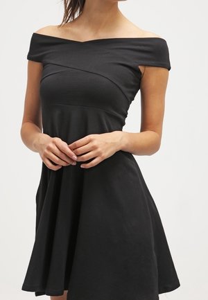 Femme portant une robe noire décolletée avec un corsage ajusté et une jupe évasée, les mains délicatement jointes devant elle.