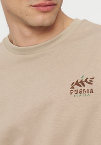 Μπεζ μπλούζα με λαιμόκοψη, που φοράει ένα άτομο, με μικρό καφέ και πράσινο λογότυπο φύλλου και το κείμενο "Puglia Italia" στο στήθος.