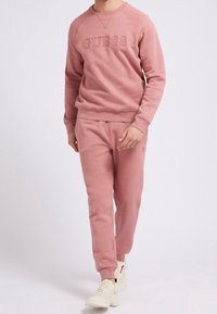 Lyserød bomulds-sweatshirt med rund halsudskæring og præget "GUESS"-logo, parret med matchende lyserøde joggingbukser. Har ribbede manchetter og linning.