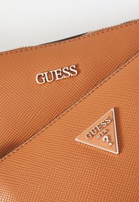 Ruskea teksturoitu materiaali-salkku, jossa on hopeinen "GUESS"-logo, kolmiomainen "GUESS USA" -symboli ja kontrastiväriset ompeleet reunoilla.