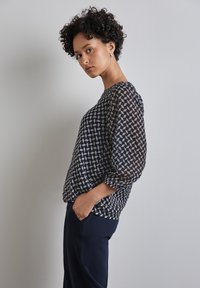 Blouse marine avec un motif géométrique blanc, en tissu transparent, et manches trois-quarts. Portée avec un pantalon marine, équipé de poches latérales.