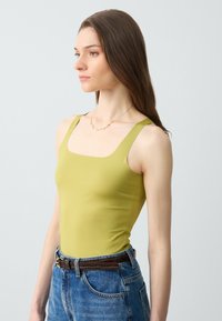 Canotta verde lime aderente con spalline larghe e scollatura quadrata, abbinata a jeans blu a vita alta e una cintura marrone.