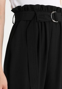 Pantalons noirs taille haute avec une ceinture élastique froncée, featuring une large ceinture avec une boucle en argent et un tissu drapé.