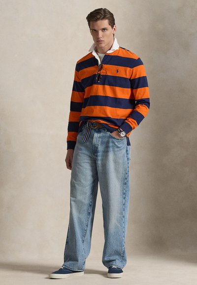 Langärmeliges Poloshirt in Orange und Marineblau gestreift, mit weißem Kragen, kombiniert mit locker sitzenden hellblauen Jeans und marineblauen Schuhen. Einfache Gürtel-Details.