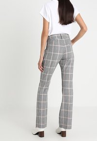 Vrouw met lang, donker haar in grijze geruite rechte broek en een witte korte mouwen top, staand met één hand op de heup, met de rug naar de kijker toegekeerd.