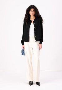 Black bomber jacket with silver buttons, white top, cream striped wide-leg trousers, and black pointed-toe shoes. Holding a blue clutch.