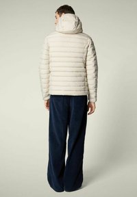 Uomo di spalle che indossa un piumino beige chiaro con cappuccio e pantaloni a gamba larga blu scuro su uno sfondo neutro.