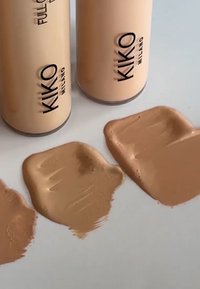 Deux bouteilles cylindriques beige étiquetées "KIKO Milano" à côté de trois échantillons de fond de teint liquide de teintes différentes sur une surface blanche.