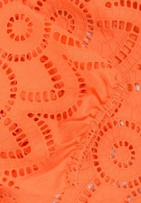 Oranje stof met een kantachtig patroon, voorzien van ronde uitsparingen, gestructureerde stitching en geplooide ontwerpelementen.