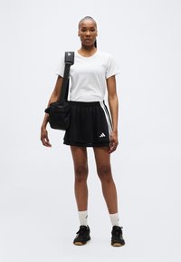 Γυναίκα με λευκό t-shirt, μαύρη αθλητική φούστα, λευκές κάλτσες και μαύρα αθλητικά παπούτσια, κρατώντας μαύρη τσάντα ώμου σε απλό φόντο.