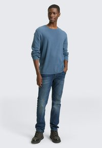 Blauer, langärmeliger, strukturierter Pullover kombiniert mit dunkelblauen Jeans und schwarzen Schnürstiefeln; Stehpose mit Händen in den Taschen vor einem hellen Hintergrund.