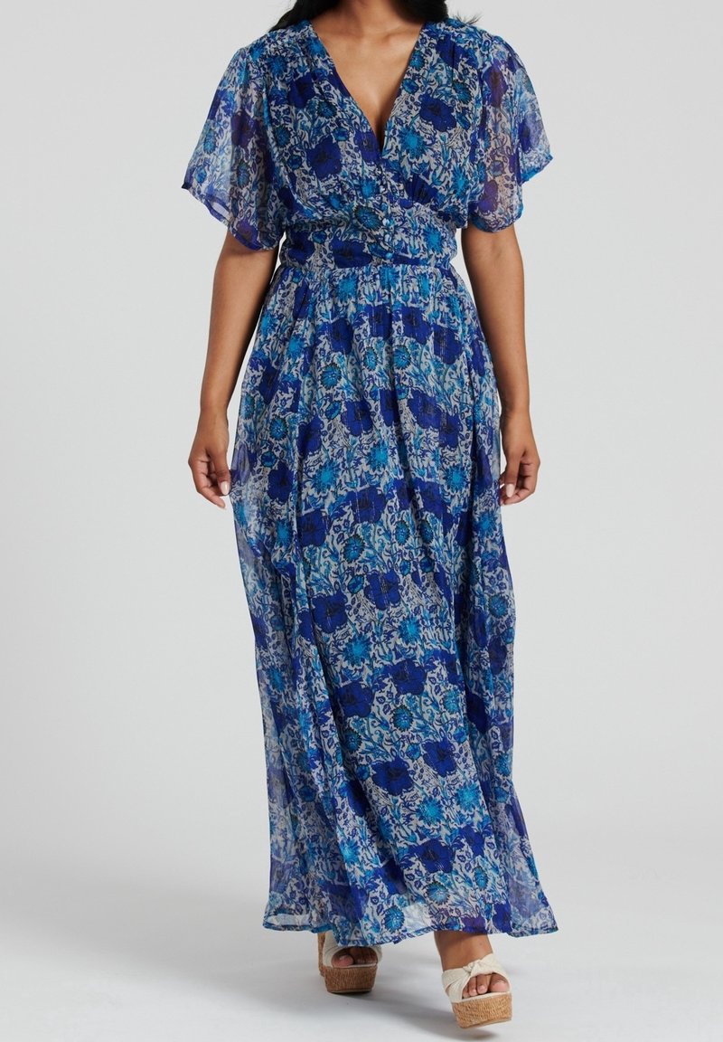 Femme portant une robe maxi bleue fleurie avec des manches courtes transparentes et un profond décolleté en V, associée à des sandales compensées beiges.