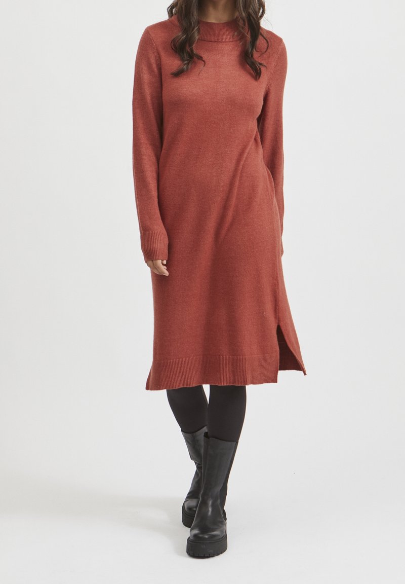 Femme portant une robe midi en maille à manches longues couleur rouille avec une fente sur le côté, des leggings noirs et des bottes noires mi-mollet sur un fond uni.