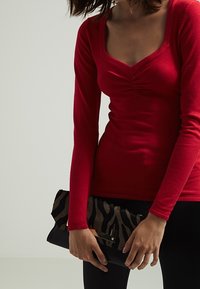 Rotes Langarmoberteil mit gerafftem Detail am Ausschnitt, kombiniert mit einer schwarz-braunen Zebra-gestreiften Clutch mit metallischem Akzent.