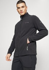 Schwarze Fleecejacke mit hohem Kragen, durchgehendem Reißverschluss, seitlichen Reißverschlusstaschen und Logodetails auf der Brust und dem Ärmel. Glatte Textur.