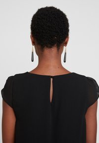 Femme aux cheveux courts et bouclés portant un haut noir avec fermeture à boutons et de longues boucles d'oreilles pendantes noires et dorées, vue de dos sur fond gris.