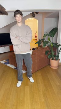 Giacca zip beige chiaro, camicia bianca e jeans grigio scuro; in piedi su un pavimento di legno vicino a un divano in velluto corduroy marrone e a una grande pianta in vaso.