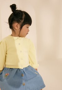 Gul cardigan med små blomsterknapper kombineret med nederdel i denim med broderede blomster på lommerne. Set fra siden.