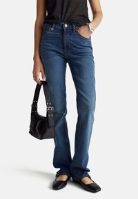 Jeans de mezclilla azules con un ligero acampanado, que presentan un diseño clásico de 5 bolsillos, costuras sutiles y una cintura de altura media. Combinados con un bolso negro.