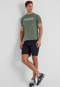 Green cotton t-shirt with white "TOG24" text, paired with navy cargo shorts and blue New Balance sneakers with gray accents.