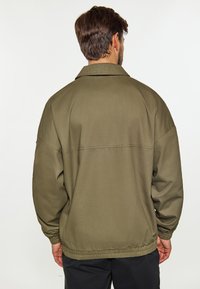 Olivengrøn jakke med en afslappet pasform, præget af en klassisk krave, elastiske manchetter og en talje. Fremstillet af glat, mat stof.