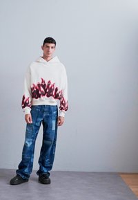 Hanorac alb cu grafică de flăcări roșii la margine, purtat cu blugi albaștri cu diverse modele imprimate, completat cu adidași negri.