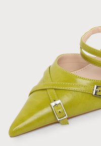 Chaussure en cuir verte à bout pointu avec des brides croisées et une boucle argentée, vue de dessus sur un fond clair.
