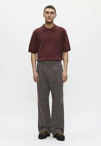 Young man standing wearing a maroon short-sleeve polo, gray wide-leg trousers, and black shoes against a plain white background.