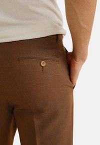 Pantaloni marroni realizzati in tessuto con una texture liscia. Presentano una tasca posteriore con chiusura a bottone e un piccolo logo del marchio.