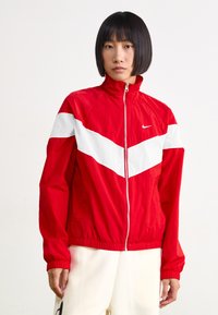 Rød zip-up vindjakke med hvit chevron-design, elastiske mansjetter og midjebånd. Laget av lettvektsstoff, med en liten logo på brystet.