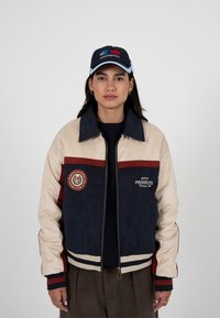 Femme portant une veste zippée bleu marine, beige et rouge avec des écussons de course et une casquette bleu marine assortie avec des logos, sur un fond uni.