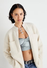 Krémová bomber bunda ze směsi vlny s dvěma předními kapsami, nošená přes stříbrný saténový crop top, spárovaná s džíny s vysokým pasem.