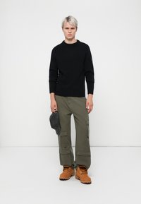 Pulover tricotat negru, pantaloni cargo măslinii, cizme maro și o șapcă gri ținută în mână. Design simplu și croială lejeră.