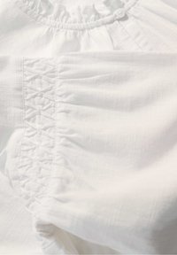 Weißer Kleidungsärmel mit elastischem Raffungdetail und einem kleinen Knopf in der Nähe des gerafften Ausschnitts, der die Stofftextur zeigt.