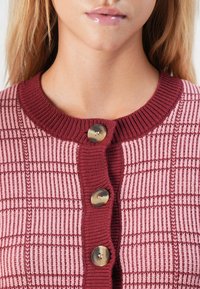 Femme portant un cardigan en tricot à motifs de carreaux roses et bourgognes avec trois grands boutons, mettant en valeur le cou et les lèvres.