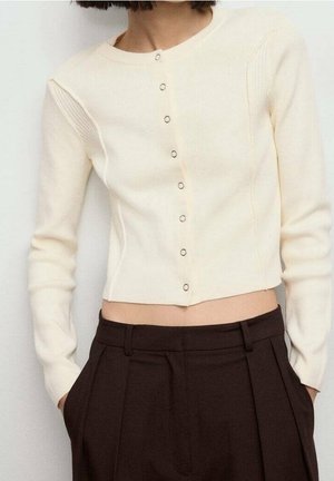 Femme portant un cardigan court couleur crème avec de petits boutons et un pantalon plissé marron foncé taille haute, les mains dans les poches.